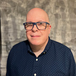 Headshot of Jeff, a bald man wearing blue glasses and a dark button-up shirt, smiling slightly against a neutral patterned gray background.
